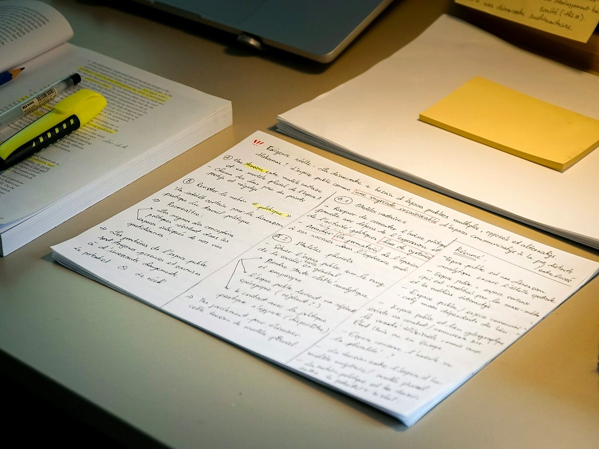 A research workspace with academic papers, a highlighted textbook, handwritten notes and a laptop